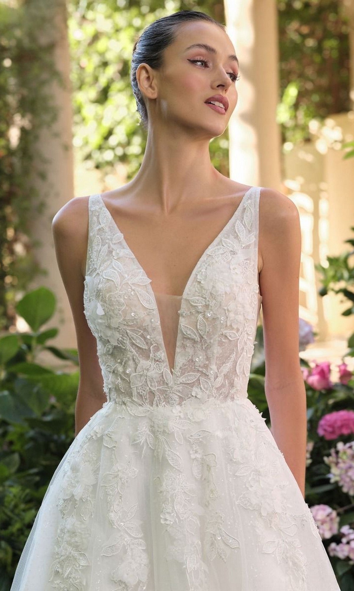 A woman stands in a garden wearing the Andrea and Leo WL064, a long wedding dress featuring an elegant A-line silhouette with a sleeveless white lace bodice and delicate appliqués.